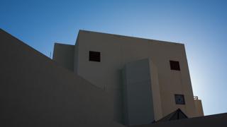 A close up image of Workman Center, looking up. The sun is behind the building and the sky above the building is cloudless and light blue.